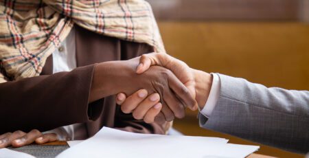 Close-up of two business people, one wearing a plaid scarf, shaking hands over papers on a desk to finalize a deal or partnership.