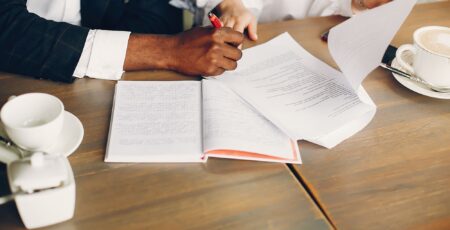 Two business partners sitting at a cafe table, one signing a document in a notebook, with coffee cups nearby.