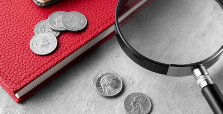 Close-up shot of a magnifying glass and coins on a red textured notebook, symbolizing financial analysis or auditing.
