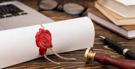 A rolled diploma with a red wax seal on a dark wooden desk, next to a sealing stamp, pen, laptop, and stacked books.