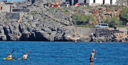 A man paddleboarding and two people kayaking on deep blue water near a rocky coastline with a white house on the cliff.